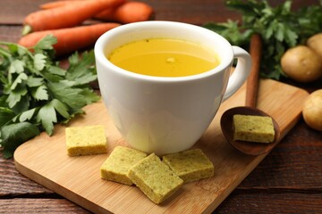 Aromatic bouillon cubes, broth, parsley and vegetables on wooden table, closeup