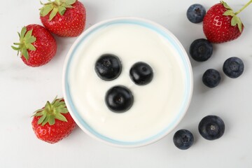 Tasty yoghurt with blueberries and strawberries in bowl on white marble table, flat lay