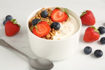 Tasty yogurt with granola, berries and spoon on white marble table, closeup