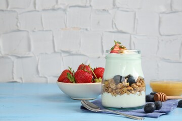 Tasty yogurt with granola, berries, honey and spoon on light blue wooden table, space for text