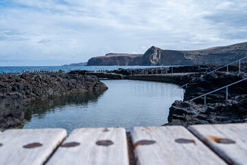 A wooden bench overlooking a body of water. The water is calm and clear, and there are rocks in the...