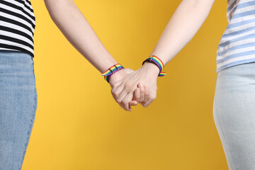 LGBT concept. Women in rainbow wristbands holding hands on yellow background, closeup