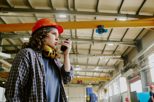 Young adult Caucasian woman wearing hard hat using walkie talkie while standing in industrial warehouse, communicating instructions with focused expression, machinery visible in background