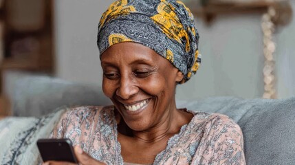 smiling middle aged african woman with traditional head turban sitting on couch at home using smartphone beautiful african american woman with typical headscarf scrolling through internet on phone no