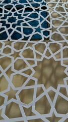 Intricate geometric pattern of white metal casting shadows against beige facade and blue tinted window