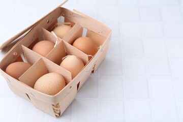 Raw chicken eggs in wooden egg tray on white tiled table, closeup. Space for text
