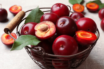 Whole, cut cherry plums, green leaves and metal basket on grey textured table, closeup