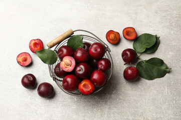 Ripe cherry plums, green leaves and metal basket on grey textured table, flat lay