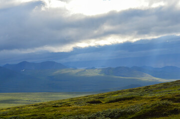A dramatic Alaskan landscape with rolling green hills, distant mountains, and sunbeams breaking through heavy clouds