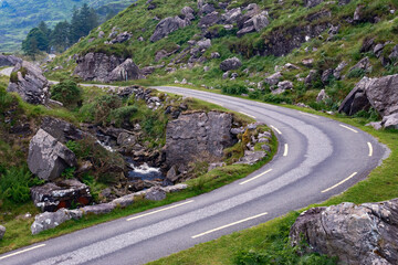 An asphalt road winds through a lush, rocky mountain pass. A small stream flows alongside the road in the Gap of Dunloe in County Kerry, Ireland. © Oleksii