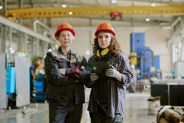 Caucasian middle aged woman and Caucasian young adult woman standing in industrial factory wearing...