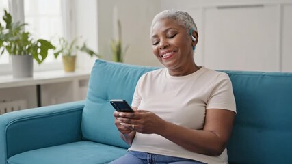 African American woman about sixty with blue hearing aid smiling and talking while using smartphone, white t-shirt, silver hair, turquoise sofa, cozy modern room, daylight, plants - Powered by Adobe