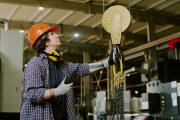 Young adult Caucasian woman operating industrial crane in factory, wearing protective helmet and gloves, holding remote control, looking upward while monitoring heavy machinery