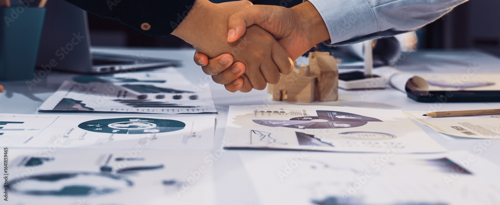 Poster a close-up of hands shaking over business strategy documents featuring graphs and charts, symbolizin