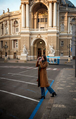 Attractive woman in coat taking a photo with film camera of old building of 18th century in Europe.