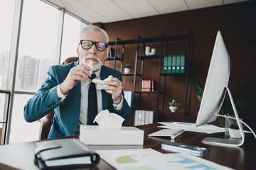 Senior businessman in office setting holding tissues while working at desk with computer...