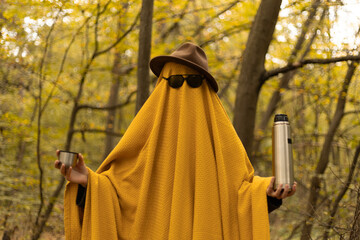 A man in a mustard-colored sheet, hat, and glasses, like a ghost in an autumn forest, offers to...