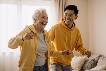Happy caucasian elderly woman and asian man dancing together in a cozy living room wearing yellow hoodies