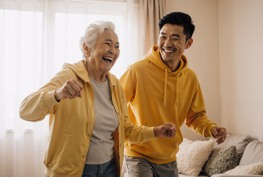 Happy caucasian elderly woman and asian man dancing together in a cozy living room wearing yellow hoodies