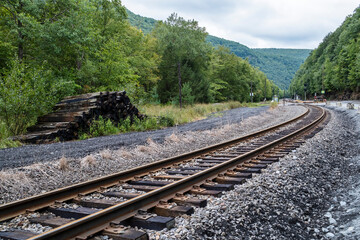 Fototapeta premium A serene view of a winding railway track surrounded by lush green foliage and mountains, with stacked wooden logs nearby, creating a tranquil atmosphere.