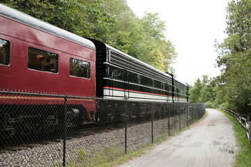 A vintage train passes by a scenic pathway, surrounded by lush greenery and a chain-link fence, evoking a sense of nostalgia and adventure.
