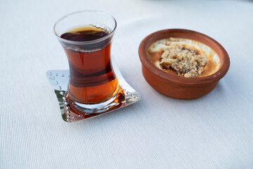 A glass of tea served on a decorative plate alongside a clay bowl filled with a sweet dessert,...