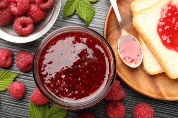 Sweet raspberry jam in glass jar, berries and bread slices on dark gray wooden table, flat lay
