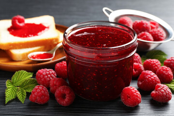 Sweet raspberry jam in glass jar, berries and bread slices on dark gray wooden table, closeup