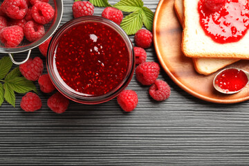 Sweet raspberry jam in glass jar, berries and bread slices on dark gray wooden table, flat lay