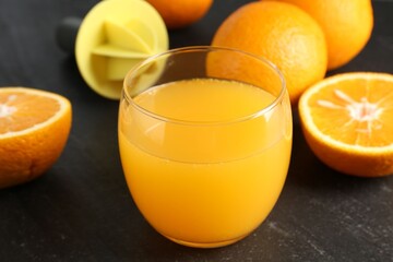 Fresh orange juice in glass, fruits and juicer on black table, closeup