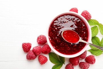 Sweet raspberry jam in bowl, berries and green leaves on white wooden table, flat lay. Space for text
