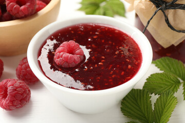 Sweet raspberry jam, berries and green leaves on white wooden table, closeup