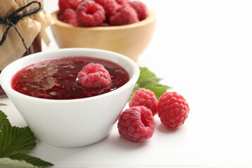 Sweet raspberry jam, berries and green leaves on white wooden table, closeup. Space for text