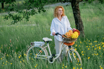 Red-haired young woman enjoying summer evening in nature with aesthetic bicycle. Basket, flowers.