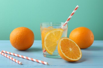 Sparkling water with orange slices in glass, fresh fruits and straws on light blue wooden table against turquoise background, closeup