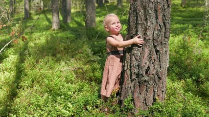 Little blonde girl wearing dress hugs a large pine tree trunk in a lush green forest on a sunny summer day, expressing love for nature and environmental awareness
