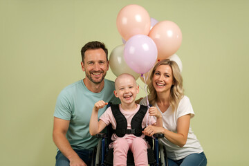 Happy family celebrating with a child cancer patient. Laughing bald boy in a wheelchair with his parents and balloons. Joy and recovery concept.