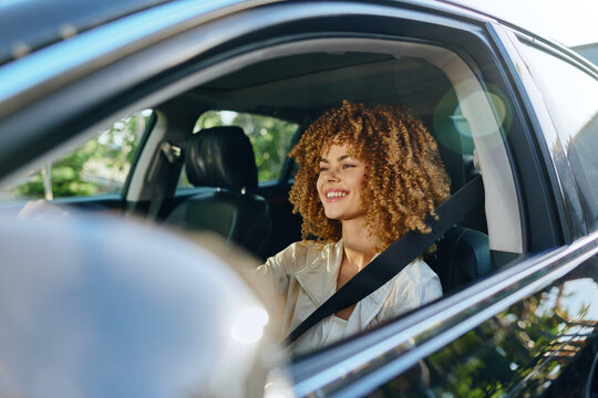 Smiling woman driving car fastened with seatbelt, curly hair, casual clothing, daylight, safe urban travel, joyful expression, natural light.