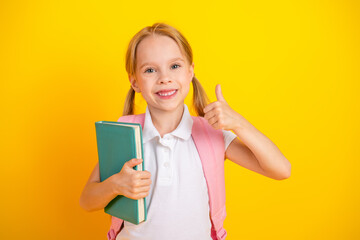 Happy schoolgirl with backpack holding a book and showing a thumbs up gesture with a bright yellow background