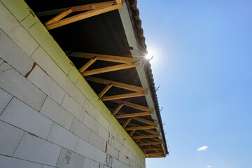 Looking up wooden roof construction at new house building site, bare wall near, sun shines on clear sky