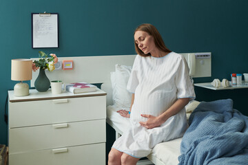 Caucasian pregnant young adult woman sitting on hospital bed holding belly with eyes closed,...