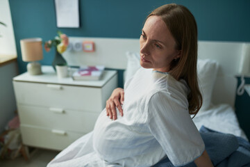 Obraz premium Caucasian pregnant young adult woman sitting on hospital bed holding belly, gazing thoughtfully to side, showing anticipation and calm expression in medical setting