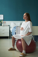 Caucasian pregnant young adult woman sitting on exercise ball holding belly breathing deeply in...
