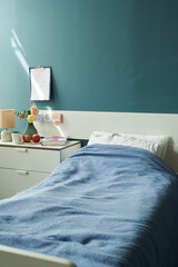 Hospital bed with blue blanket standing in medical room, bedside table holding lamp, flowers,...