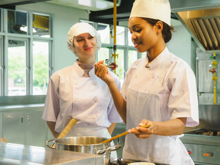 Professional female two chefs tasting food in culinary training education modern kitchen wearing white uniforms working together in clean, One chef tasting soup from wooden spoon while other watches