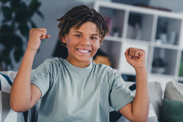Happy boy raising arms in joy while sitting at home in a comfortable living room, dressed in a casual t-shirt
