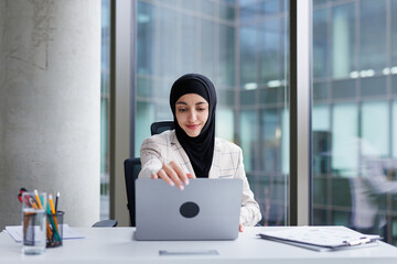 Office employee in hijab opening laptop after coming to workplace