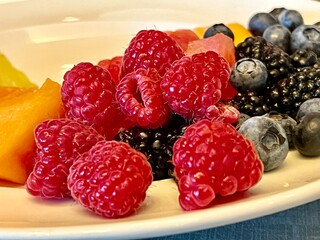 Close-up of fresh raspberries, blueberries, blackberries, and melon pieces on a white plate,...