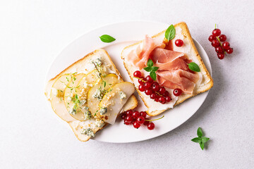 Tasty homemade toasts with jamon, cream cheese, red currant and with cheese, pear, gorgonzola and nuts onwhite ceramic plate on light stone background top view.