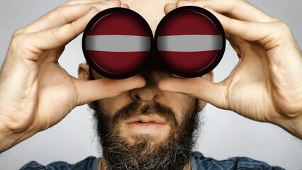 The Latvian flag is reflected in the lenses of binoculars through which a bearded man looks